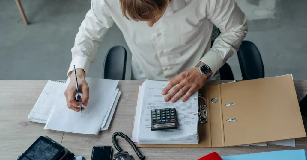 Business owner reviewing and organizing business sale documents at desk with calculator and paperwork.