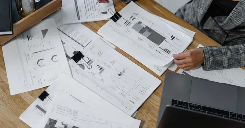 Designer reviewing business sale documents and wireframes on wooden desk with laptop.