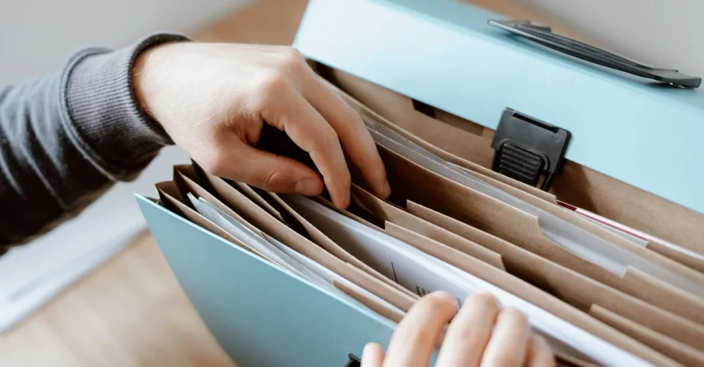 Hands organizing labeled folders in filing box with estate planning documents and legal paperwork organized by tabs.