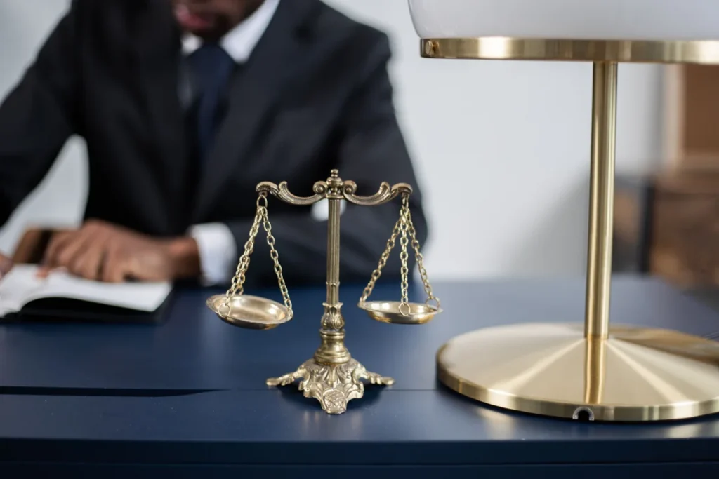 Close-up of brass scales of justice on a desk with a brass lamp and a blurred person in a suit reading a book in the background..