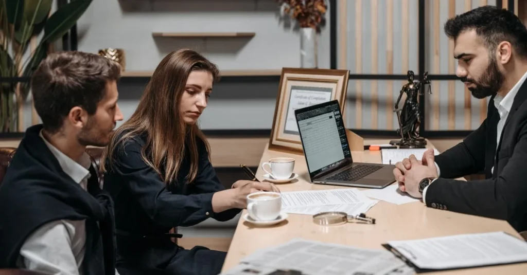 Three professionals in business attire discussing documents at a meeting table with laptop and coffee cups.